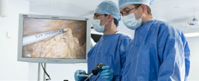Two surgeons in surgical scrubs and masks are practicing laparoscopic surgery. In the background a monitor displays their instruments and the procedure they are performing.